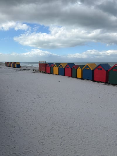 The image shows a row of colorful beach huts in Muizenberg, South Africa. The huts are painted in various colors including green, red, blue, and yellow. The beach is sandy and stretches towards the horizon. The sky is partly cloudy with a mix of gray and white clouds, with patches of blue sky visible. The ocean is visible in the background.