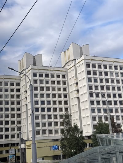 The image shows a tall, white, multi-story building with a grid of square windows. The building has a modern architectural style with decorative elements at the top. A street light pole is prominently positioned in the foreground, obscuring part of the building. Power lines are visible crossing the sky above the building. Trees are growing in front of the building, and a glass structure, possibly a bus stop shelter, is in the lower right corner. The sky is partly cloudy. The location is identified as Сеніцкі сельскі Савет, Belarus.