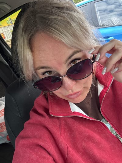 The image shows a close-up, self-taken portrait of a blonde woman with sunglasses, wearing a red zippered jacket over a brown shirt, inside a vehicle. The woman is holding her sunglasses with her right hand, revealing her eye and part of her face. Her nails are painted in a light color with silver glitter accents. The background shows the interior of a car, with a blue car visible through the window. The scene is likely in the city of Chastyye, Russia, as indicated by text on a yellow sign in the background which reads "ЧАСТЫЕ" (Chastyye).