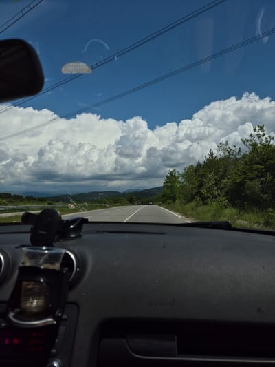 The image provides a daytime forward-facing view from inside a vehicle traveling in the area of Ponor, Bulgaria. The lower half of the frame displays the vehicle's dark gray dashboard. On the left side, a black car phone holder is mounted, and below it, a black cupholder, attached to an air vent, contains a translucent plastic cup with a small amount of residual liquid. A portion of the rearview mirror is visible in the upper left, reflecting interior elements of the vehicle. The windshield shows various reflections and smudges, including distinct horizontal power lines, a semicircular arc, and a grid-like pattern, superimposed against the sky.
The vehicle is on an asphalt road with a solid white line marking its right edge and a broken white line indicating lane separation. A silver metal guardrail runs parallel to the road on the left, beyond which is a vegetated slope. The immediate roadside is covered with green grass, trees, and shrubs.
In the mid-ground and background, the road extends into a rural and mountainous landscape. Distant green hills are densely covered with foliage, and other vehicles are faintly visible on the road ahead. The sky transitions from clear blue in the upper regions to being predominantly filled with large, white cumulus clouds, some of which appear darker and more diffused.