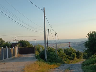 The image depicts a view from a hillside overlooking a coastal city and an airport. In the foreground, a dirt road curves to the right. To the left, a grey metal fence with decorative spherical lights runs along a concrete wall. A large dark metal gate is visible beyond the fence, leading into a property with a red-roofed building partially obscured by trees. Several utility poles with wires traverse the sky. In the middle ground, lush green vegetation and trees cover the hillside. In the background, an airport runway with red and white markings is visible, alongside the city buildings and the sea. The sky is clear and a pale blue, transitioning to a light yellow near the horizon, suggesting it is either early morning or late evening. The overall scene is tranquil and serene. The location is Chereshnya, Russia.