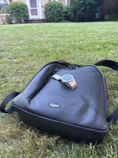 A close-up, low-angle shot shows a black pebbled leather backpack lying on a patch of green grass in Shaker Heights, United States of America. A silver wristwatch with a blue face rests on top of the backpack, near a zippered compartment. The Lacoste logo, a silver crocodile, is visible on the front of the backpack. The background is out of focus, showing a house with bushes and a white door.