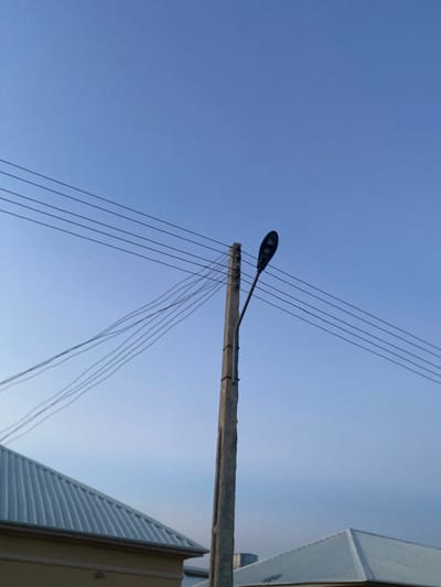 The image captures a low-angle view of a clear blue sky, with power lines and a street lamp visible against it. The power lines are numerous and run diagonally across the frame, appearing to be suspended from a concrete pole. A dark, modern-looking street lamp is attached to this pole. In the foreground and along the right edge of the image, a portion of a light-colored building with a corrugated roof is visible, suggesting an urban or semi-urban setting. The clear sky and bright light indicate it is daytime, likely morning or afternoon. There are no people or specific activities depicted. The scene is devoid of any visible text. The location context provided is Dumnga, Nigeria.
