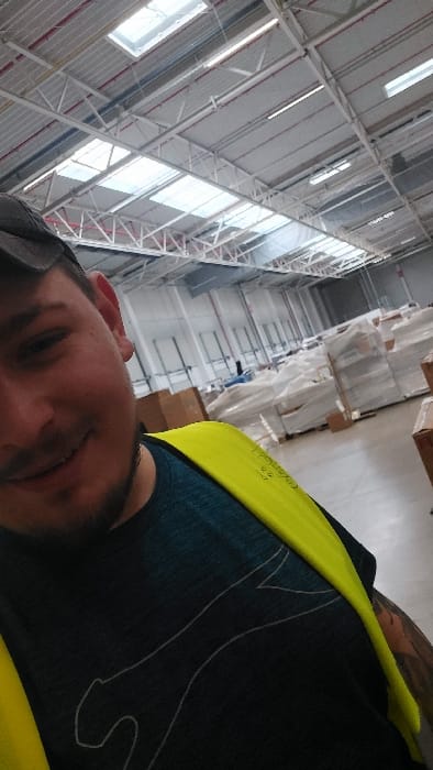 The image displays a male individual capturing a selfie within a large industrial warehouse located in Senec, Slovakia. The man, positioned in the foreground to the left, is smiling and wears a dark t-shirt with a light-colored stylized animal silhouette, a bright yellow high-visibility vest, and a dark baseball cap. A tattoo is partially visible on his right arm. The background reveals an extensive warehouse interior characterized by a high ceiling composed of white metal trusses, multiple rectangular skylights providing natural illumination, linear fluorescent light fixtures, and exposed red and blue piping. The floor appears to be concrete. Stacked pallets laden with goods, predominantly wrapped in white or clear plastic, are visible throughout the midground and background, interspersed with several brown cardboard boxes. Some indistinct shapes, potentially other persons or machinery, are discernible further within the warehouse space.