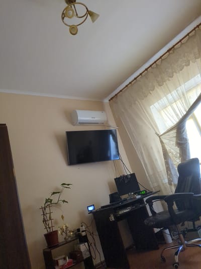 The image depicts an indoor scene, likely a room in Vilne, Ukraine. In the upper portion of the image, a ceiling light fixture with three arms and glass shades is visible against a white ceiling. Below the ceiling, a beige wall is present. Mounted on the wall is a white air conditioning unit, and beneath it, a black flat-screen television is displayed.

To the right of the television, a black desk is set up, featuring a computer monitor displaying the time "11:59", a computer tower, and various cables. An office chair with a black leather seat and chrome legs is positioned in front of the desk. A light-colored curtain with an ornate pattern hangs over a window to the right of the desk.

To the left of the desk and below the television, a potted plant with green leaves supported by a wooden trellis is situated on a shelf unit. This shelf unit also holds decorative items and power strips with multiple plugs. The overall impression is that of a living space or office area.