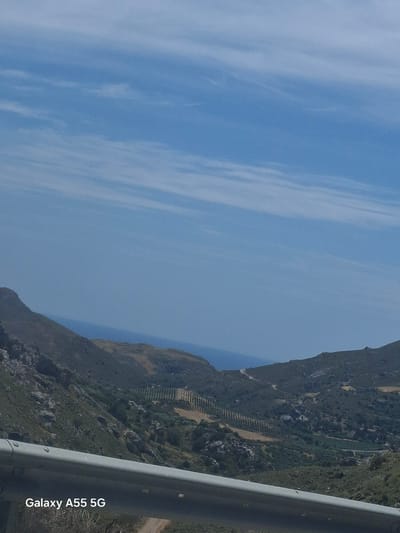 The image shows a landscape with a mountain ridge covered in vegetation on the right side of the frame. A section of a gray metal guardrail is visible in the lower right corner. The sky, displaying alternating shades of blue and white, fills the majority of the image to the left. The photo appears to have been taken from a vehicle. The watermark "Galaxy A55 5G" is present in the lower right corner.

The location is in the city of Frati, Greece.