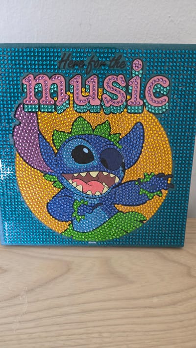 The image depicts a diamond art piece of the Disney character Stitch in front of a yellow and green circular background, with the word "Stitch" written vertically in pink. The artwork is made of small, colorful, shiny rhinestones affixed to a teal-colored canvas. The background consists of blue rhinestones. The image appears to be photographed indoors, likely attached to either a wall or furniture, with a white wall partially visible on the left and a wooden surface on the right. There is no indication of Zirndorf, Germany in the image.