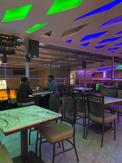 This image captures an indoor dining or lounge area, likely in the evening or night, indicated by the artificial lighting. The scene features modern and traditional design elements.
Two men are visible within the establishment. One man, wearing a light-colored shirt, dark cap, and glasses, stands leaning over a marble-patterned table, appearing to focus on something on its surface. Another man, dressed in a dark jacket, is seated at an adjacent table.
The space is furnished with numerous tables featuring white and grey marble-patterned tops, paired with chairs that have ornate, possibly cast-iron, backs displaying a Greek key design, and light brown or beige upholstered seats.
A distinctive feature of the setting is the ceiling, which boasts geometric cutouts illuminated with vibrant green and blue lights, creating a modern and dynamic ambiance. Other artificial lights in the background emit purple and pink hues. A black speaker box, clearly labeled "NEXO," is mounted on a stand or pole, suggesting the venue might host events with music or performances. The floor is light-colored, possibly tiled, and reflective surfaces or glass panels are seen in the background, contributing to the contemporary feel.
The overall atmosphere appears quiet, with only the two individuals present in the visible section of the room. There are no direct visual cues in the image itself to confirm the location as Kimorori, Kenya, beyond the provided context.