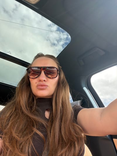 A woman is taking a selfie in a car, with the roof of the car open to the sky. The woman has long, brown hair and is wearing sunglasses and a brown shirt with a cutout. Her lips are pursed. The car interior is visible, including a tan seat and black trim. The sky is cloudy. The image appears to be taken during the day.