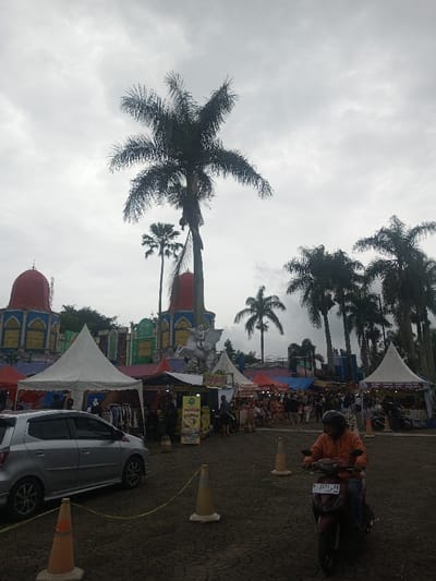 The image portrays an outdoor market or event in Suddepalle, India, under an overcast, grey sky. The ground in the foreground consists of dark, cracked earth. Three orange and white traffic cones are visible, with yellow caution tape extending from a cone on the lower left.
On the left, a silver five-door hatchback is parked, facing left. On the right, a person wearing an orange jacket and a dark helmet rides a dark red scooter, license plate F 2271 TE, directly towards the viewer.
The midground is dominated by an active market featuring numerous white pointed-roof tents and various colored stalls. Approximately twenty to thirty individuals are dispersed throughout the market area, engaged in activities such as standing, walking, and congregating. Merchandise, including clothing, is visible at some stalls, and a sign depicting a chicken is present at one stall.
In the background, behind the market, distinctive structures are present. A prominent white statue of a winged horse carrying a rider is centrally located. To the left, two ornate buildings with red dome roofs and intricate blue and yellow patterned bases stand, accompanied by other green and multicolored architectural elements. Multiple tall palm trees are scattered across the scene, with some towering significantly above the structures and market tents.