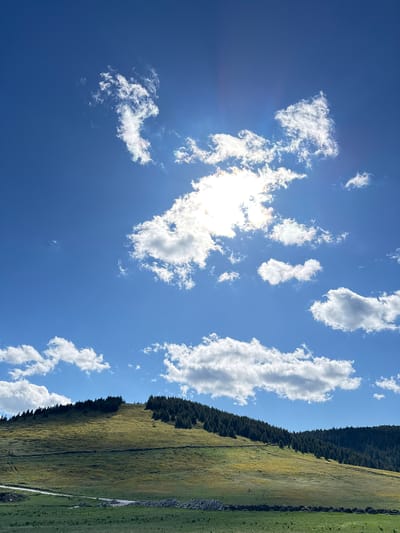 The image depicts a sunny day with a clear blue sky dotted with fluffy white clouds. The sun is partially obscured by clouds, casting rays of light downwards. In the foreground, there is a grassy hill with a scattering of trees along its crest. A dirt road winds its way up the side of the hill. The overall scene is tranquil and natural, evoking a sense of peace and serenity. The image does not contain any information about the city of Gela, Bulgaria.