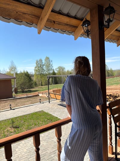 A person is standing on a porch in Kamennoye, Russia. The person is wearing a blue striped shirt and pants. The porch is made of wood and has a gray tiled floor. There is a swing set in the yard. The sky is blue and there are some trees in the background.