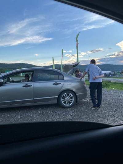 In the city of Inzer, Russia, a silver sedan is parked on a gravel lot next to a road. The rear trunk of the car is open. A woman with sunglasses is seated in the driver's seat of the car. Two men are standing behind the car, with one of them looking into the trunk. The background shows a road, buildings, and forested hills under a cloudy sky.