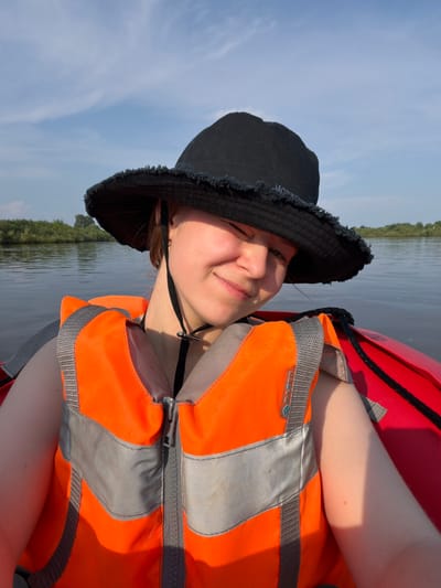 A young woman with light skin and auburn hair is wearing a black bucket hat and an orange life vest. She is in a red kayak on a body of water, possibly a river or lake, with trees lining the distant shore. The sky is partly cloudy and blue. She is looking at the camera and smiling. The scene appears to be outdoors in a natural environment. The location is the city of Kalino, Russia.