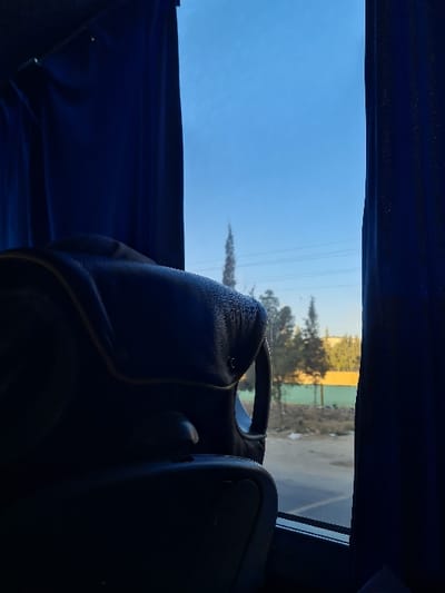 The image displays an exterior view from inside a vehicle, likely a bus, looking out a window in Ashrafia, Syria. Dark blue curtains frame the window on both the left and right sides. The foreground on the left is occupied by the top portion of a dark, possibly leather-upholstered seatback, featuring a headrest and an integrated handle, which partially obstructs the lower window. Outside the window, a dirt or gravel roadside with sparse dry vegetation and scattered debris is visible. Beyond this roadside, a landscape with multiple green-leafed trees is present, including a distinctively tall and slender coniferous tree positioned against the sky. Horizontal utility lines traverse the mid-ground, visible across the clear blue sky above and behind the trees. In the background to the right, a building with a prominent yellow wall is partially visible through the trees, adjacent to what appears to be a green barrier or fence. The exterior scene is brightly illuminated by natural daylight, contrasting with the shadowed interior of the vehicle. No individuals are visible.