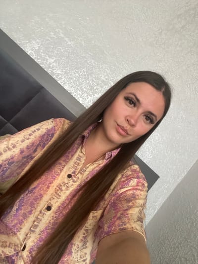 A young woman with long, dark brown hair and prominent eyelashes poses for a selfie. She is wearing a button-up shirt with a gold and purple abstract pattern. The background appears to be a bedroom with a textured white wall and a dark upholstered headboard. There is no identifiable information within the image to suggest the location is Chalda, Russia.