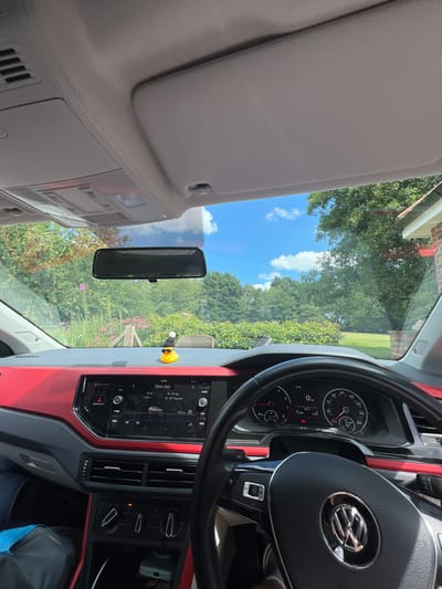 The inside of a Volkswagen car is visible from the driver's perspective. The dashboard, steering wheel with the VW logo, and the rearview mirror are prominent. A sun visor is folded down on the left. Outside the windshield, trees and a blue sky with clouds can be seen. A small yellow rubber duck is on the dashboard.
