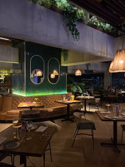 This image depicts the interior of a dimly lit restaurant or bar, identified as "Peregrina," located in Floridablanca, Colombia, during nighttime hours.
The scene focuses on a main dining area featuring a long, curved, tufted brown leather booth set against a prominent wall. This wall is clad in dark green, small-scale, possibly fish-scale pattern tiles, illuminated at its base by a strip of green LED lighting. Embedded within this green tiled wall are two vertical, oval-shaped mirrors with thin metallic frames. Above this section, a substantial concrete beam forms part of the ceiling structure, featuring planter boxes filled with artificial or live green foliage, including hanging vines.
The flooring consists of light-toned wooden or wood-effect planks. Numerous dark wooden tables with black metal bases are arranged across the floor area. These tables are predominantly set for dining, displaying white plates, cutlery, clear wine glasses, and water glasses. Several tables also feature small, portable, illuminated table lamps and circular red or pink placemats.
Lighting is primarily ambient, originating from several large, textured pendant lamps with woven shades suspended from the ceiling, small table lamps, and accent lighting on the green wall.
In the background, to the left, a bar area is visible with bottles on shelves and glassware. To the right, through large windows, a panoramic nighttime view of a city skyline with illuminated buildings is discernible.
At least two individuals are faintly visible seated at a table near the windows in the distant background, appearing as patrons. No direct interactions or specific actions are clearly distinguishable. The environment appears clean and well-maintained, set for dinner service.