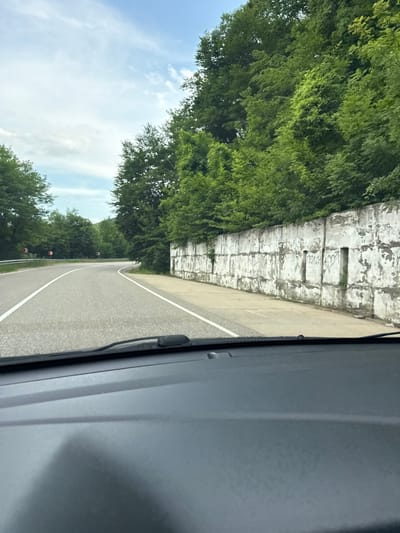 A view from inside a vehicle shows a curved asphalt road flanked by a concrete wall on the right and trees on both sides. The concrete wall is weathered and discolored, with some graffiti visible, including the word "ПОЛИНА" and a heart symbol. Beyond the wall, a dense forest of green trees rises up a slope. The road has white lane markings, and a few traffic signs are visible in the distance on the left, indicating sharp curves. The sky above is partly cloudy. The interior of the vehicle, including the dashboard and windshield wipers, is visible in the foreground. The scene appears to be a road in a rural or mountainous area, possibly near the city of Kurinskaya, Russia, given the context provided.