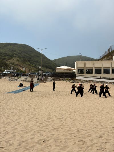 The image shows Praia das Maçãs, a beach located in Colares, Portugal. The scene captures a group of people practicing martial arts on the sandy beach. The group consists of several individuals, mostly young women, dressed in black martial arts uniforms and white or pink headbands, performing various stances and movements. In the background, there is a light-colored building with large windows, a white umbrella, and some parked vehicles, including a camper van. Further back, a lush green hillside forms the backdrop, with a road and a fence visible along its slope. The sky is overcast with a light haze. The sand on the beach has a textured surface with footprints and small debris. A blue mat is laid out on the sand in the foreground, with two people standing on it. Another individual is standing to the right of the mat, facing away from the camera.