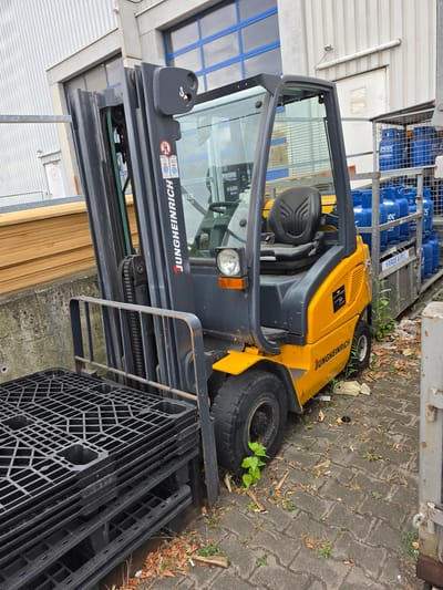 A yellow and gray Jungheinrich forklift is parked on a paved surface next to a stack of black plastic pallets. The forklift has a safety cage and a single seat. In the background, there is a metal building with blue-tinted windows and a stack of wooden boards. To the right of the forklift, a metal cage holds several blue propane tanks. The ground is covered with some dry leaves and small plants. The overall scene suggests an industrial or storage area, possibly located in Zeppelinheim, Germany, given the context. There are no people visible in the image.