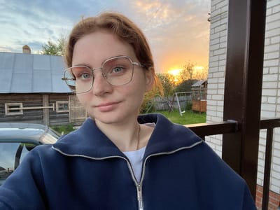 A young person, appearing to be female, takes a selfie outdoors, captured from an upward angle and presented upside down. She has light-colored hair, visible freckles, and wears silver-rimmed glasses. Her eyes are light-colored, possibly blue or green, and she has a neutral to slightly pensive expression. She is wearing a dark blue zip-up hoodie over a white t-shirt, with a thin gold chain necklace visible around her neck.
The background, when viewed correctly (rotated 180 degrees), shows an outdoor setting during what appears to be a sunrise or sunset, indicated by the warm, golden light on the horizon on the left side of the frame. The sky is partially cloudy, with soft light. To her left, there's a dark brown metal structure, possibly a railing or support beam, next to a white brick wall. Further in the background, there's green foliage, suggesting trees or bushes. On her right, a portion of a vehicle, possibly a car, is visible, reflecting the sky. Beyond that, a rustic wooden building with small windows can be seen. The overall setting suggests a residential or semi-rural area. There is no visible text in the image.