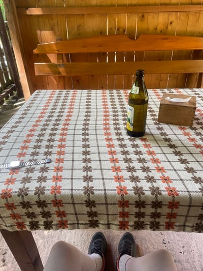 The image captures an outdoor or semi-enclosed setting, likely a patio or gazebo, viewed from above. The primary subject is a table adorned with a light-colored tablecloth patterned with alternating rows of brown and orange floral or geometric motifs.
On the table, a dark green glass bottle with a white and yellow label is visible. Partial Cyrillic text, including "КИЙ" and "Лимон" (Lemon), suggests it's a lemon-flavored beverage from a Ukrainian brand like Obolon. A wooden tissue box containing a white tissue sits near the bottle. Toward the bottom of the frame, a silver digital watch, possibly a Casio F-91W, rests on the tablecloth.
To the right of the table, the lower legs and feet of a person are seen, clad in light-colored leggings or pants, white socks, and dark athletic shoes. They appear to be seated at the table. The ground beneath their feet is a concrete-like surface. The background features light wooden planks, indicative of the structure's construction.
The scene suggests a casual daytime gathering or a moment of relaxation in Kamianytsia, Ukraine. The visible sunlight and clear textures imply pleasant weather suitable for being outdoors. The bottle and tissue box indicate a pause for refreshments or a meal.