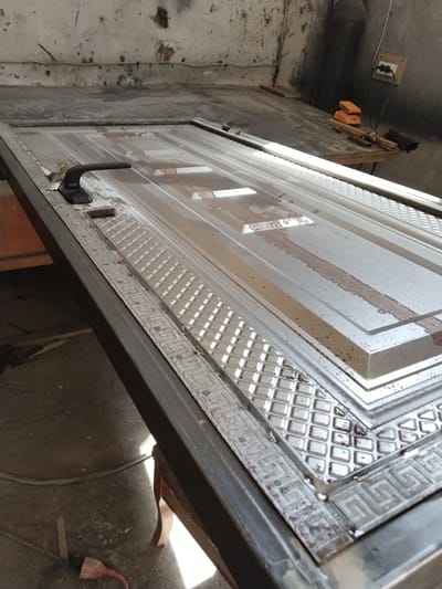 In Chalysh, Uzbekistan, a metal security door lies flat on a workbench. The door is silver and has a decorative embossed pattern. A black door handle is attached to the left side of the door, with a set of keys resting beside it. The workshop environment appears utilitarian, with tools and materials visible in the background.