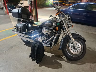 A black motorcycle is parked on a concrete surface in what appears to be an urban industrial area. The motorcycle has saddlebags and a backrest. A black backpack is on the ground next to the motorcycle. In the background, a dark blue sedan is parked, and beyond that, a yellow bus is visible. The environment suggests a commercial or service area, potentially a parking lot or loading dock. There are no people visible in the image. The lighting indicates it is either dusk or nighttime. The location, Barri De Porta, Spain, is identified.