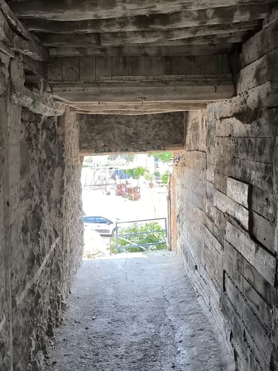 The image shows a view looking out of a rough-hewn, covered passageway, possibly a tunnel or underpass, made of stone and wood. The walls on the left and right are textured, with the left wall appearing to be rough stone and the right wall constructed of horizontal wooden planks. The ceiling is also composed of rough wooden beams and planks.
The passageway opens onto a brightly lit outdoor area. In the distance, there are glimpses of construction or renovation activity, with scaffolding, materials, and a white car parked. Greenery, likely trees and bushes, is visible. A metal railing runs along the edge of the outdoor area, suggesting a walkway or overlook.
The lighting within the passageway is dim and shadowed, contrasting with the brighter light from the outside. The ground inside the passageway is covered in a coarse, granular material, possibly gravel or dirt. The overall impression is one of an aged or unfinished structure, possibly located in an area undergoing development or maintenance.
While the image depicts a physical location, it does not contain any specific information that would definitively place it in Eğirdir, Turkey. The description provided is based solely on the visual content of the image.