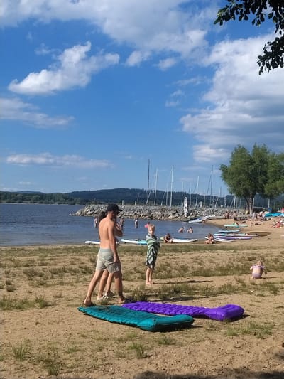 The image depicts a sunny day at a lakeside beach in Radslav, Czechia. In the foreground, two inflatable mattresses, one turquoise and one purple, lie on the sandy ground. Several people are visible near the water's edge and on the beach. A man wearing a black cap and shorts stands with his back to the camera. A child wrapped in a striped towel stands to his left, facing the viewer. Further back, various individuals are swimming or wading in the water, and some are relaxing on the sandy shore. Paddleboards and other watercraft are lined up on the beach and in the shallow water. In the background, a rocky breakwater juts into the lake, and beyond that, a dense forest covers rolling hills. Numerous sailboat masts are visible on the water, suggesting a marina or sailing club. The sky is blue with scattered white clouds.