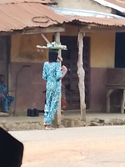 The image depicts a woman standing outside a building with a corrugated metal roof in Iseyin, Nigeria. She is wearing a long, blue patterned dress and has an object, possibly food or merchandise, balanced on her head. Her head is covered with a dark cloth. To her left, another person, partially visible, is seated and also wearing a blue patterned garment. The building appears to be a simple structure with a weathered exterior. In the foreground, there is a paved surface, possibly a street or pathway. The scene conveys a sense of everyday life and commerce in a Nigerian urban setting.