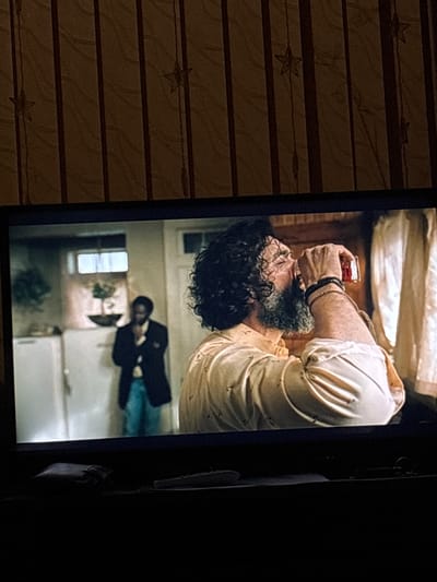 An image displays a screen, likely a television or monitor, showing a scene that is rotated 90 degrees clockwise. On the screen, a man with dark, curly hair and a full beard is prominently featured. He is wearing a light-colored, short-sleeved shirt and is shown in the process of drinking from a small, clear glass containing a reddish liquid. Dark bracelets are visible on his right wrist. In the background of the screen, a second, less distinct individual is visible, dressed in a dark top or jacket and blue pants, standing near what appears to be a white refrigerator. A window or bright light source is also present in the background of the displayed scene, suggesting an indoor setting like a kitchen or dining area.

The screen itself is set within a dimly lit room. To the left of the screen, a wall is visible, decorated with horizontal stripes and scattered star patterns, indicating the interior decor of the room where the photograph was taken. The overall lighting of the physical room is low, contrasting with the illuminated screen. No visible text is present within the image. The scene on the screen captures a moment of consumption, with the man focused on drinking. The image provides no specific time of day or weather cues beyond the general dimness of the surrounding room. The location context for this media file is Brenitsa, Bulgaria.