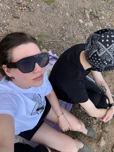 The image shows two people sitting on a purple blanket spread out on a pebbled surface, possibly a beach or riverbank. One person is wearing a white t-shirt, black shorts, and sunglasses. The other person is wearing a black t-shirt, black ripped jeans, a bandana bucket hat, and green shoes. The person in black is sitting with crossed legs, and the person in white is lying back. The location is likely near Doksha, Russia, given that was where the image was taken.