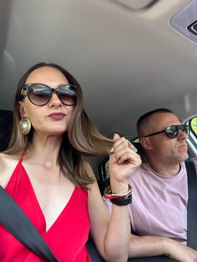 A woman and a man are inside a car. The woman, in the foreground, is wearing a red top and large, dark sunglasses. She is holding a strand of her hair. The man, in the background, is wearing a light pink t-shirt and sunglasses. He is looking to the right. The car appears to be moving, with the reflection of a road visible in the woman's sunglasses. The interior of the car, including the roof lining and seats, is visible.