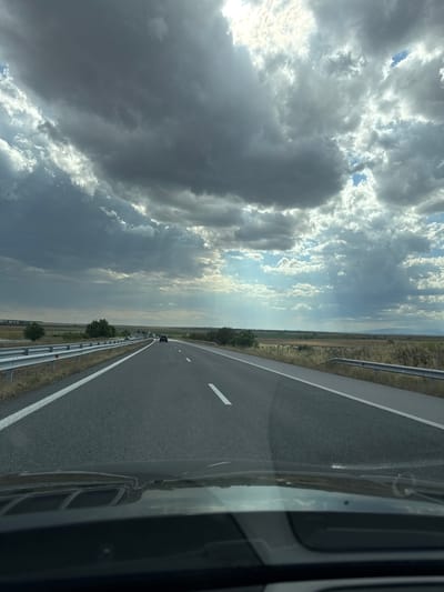 The image depicts a highway driving perspective on a cloudy day. The viewpoint is from inside a vehicle, looking forward through the windshield, which reflects the sky and the car's dashboard. The road is asphalt, with white lane markings. A silver guardrail runs along the right side of the road, separating it from a dry, grassy field. In the distance, there are trees, fields, and hills, with a hazy horizon. The sky is filled with dramatic, heavy clouds, with some shafts of sunlight breaking through. A few vehicles are visible on the highway ahead. The scene suggests a journey through a rural or semi-rural landscape. The image is located in the city of Zheleznik, Bulgaria.