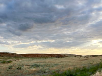 The image depicts a wide, open landscape under a dramatic sky. In the foreground, dry, grassy terrain with patches of green vegetation stretches across the scene. The middle ground is dominated by a reddish-brown embankment or hill, with a rugged, eroded surface. The background features a vast sky filled with textured, grey and white clouds, through which rays of sunlight are breaking, suggesting either sunrise or sunset. The location is identified as Ильинка, Russia. There are no visible people, animals, or man-made objects in the frame. The overall impression is one of a natural, somewhat desolate, and atmospheric landscape.