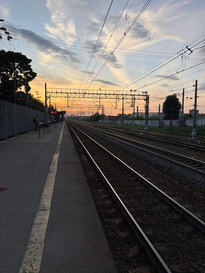 The image depicts a train station platform at sunset in Solnechnogorsk, Russia. The sky is a gradient of orange, pink, and blue, with wispy clouds. Multiple sets of train tracks run parallel, extending into the distance. Overhead, a complex network of power lines and supports crisscrosses the sky. On the platform to the left, a few people are visible, one standing near a fence and another seated at a bench under a shelter. A sign with the number "1" and Russian text is mounted on a pole. The platform is paved, with a white line marking the edge. Vegetation and buildings are visible in the background.