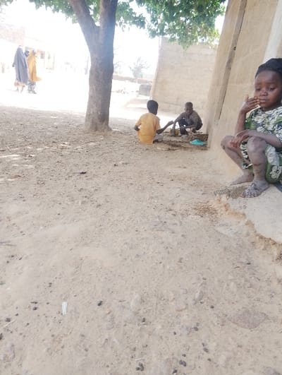 The image depicts an outdoor scene in Kanwa, Nigeria, under bright daylight conditions. The ground is composed of light brown, dry earth with scattered pebbles and minor debris.
In the foreground, to the right, a young girl, estimated to be between 5 and 7 years old, with dark braided hair, squats barefoot on the edge of a low, light-colored concrete or stone ledge. She wears a long-sleeved dress with a green, white, and black patterned fabric. Her right hand is positioned near her mouth or chin.
In the mid-ground, beneath a tree with green foliage, two young boys are seated on the ground. The boy on the left wears a light yellow or orange long-sleeved shirt and light-colored trousers, positioned with his back partially to the camera, facing towards the second boy. The second boy, wearing dark clothing, is crouched opposite the first boy, engaged with items on the ground between them. A pair of light blue open-toed sandals are visible on the ground near the second boy.
In the bright, slightly overexposed background on the left, two adult figures are walking away from the camera. One figure wears a dark, possibly blue, flowing garment, and the other wears a light yellow or tan flowing garment, including a head covering.
A light-colored building wall, constructed from blocks, is partially visible behind the squatting girl and extends into the mid-ground on the right side of the frame.