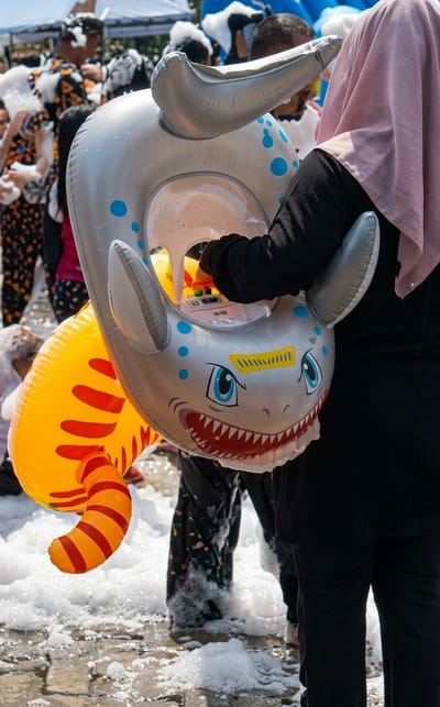 A medium shot taken outdoors depicts a person in a black long-sleeved shirt and black pants holding a gray inflatable shark toy with blue polka dots and a yellow-and-red striped tail. The person is wearing a pink hijab. They are standing on a ground covered in white foam. In the background, there are multiple people walking around. Some are wearing colorful clothing, and some appear to be covered in foam as well. It appears to be a foam party or event. The location may be Durin Simbelang, Indonesia.