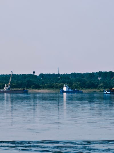 A wide, calm river flows in the foreground of the image. In the middle ground, a forested shoreline is visible, with trees of varying shades of green forming a dense canopy. A tall, dark water tower stands out against the trees. Several vessels are present on the river. On the left, a barge with a large crane is docked near the shore. Further to the right, a white and blue boat is navigating the water. On the far right, part of another barge, possibly carrying cargo, is visible. The sky above is a pale, hazy blue, suggesting it is daytime. The overall scene depicts a serene river landscape with industrial elements.