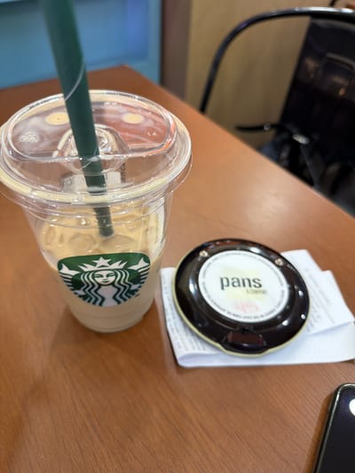 The image shows a Starbucks iced beverage with a green straw on a wooden table, along with a "pans & company" device (possibly a promotional item or ordering device) resting on a receipt. A dark-colored chair is partially visible in the background. The location is likely within Ondara, Spain, given the presence of the "pans & company" device. A mobile phone is partially visible on the upper-right corner.