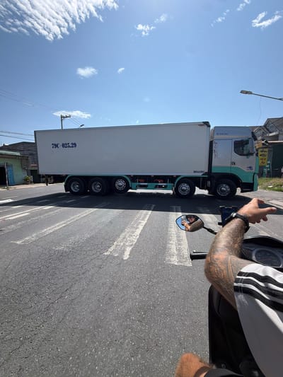 This daytime image, captured in Cam Ranh Ward, Vietnam, shows a person on a motorcycle or scooter alongside a large truck on a sunny day. The perspective is from the motorcycle rider, looking towards the truck.
The primary subject is a person, whose right arm, hand, and upper thigh are visible, indicating they are operating a motorcycle or scooter. The person wears a grey and black striped shirt (possibly Adidas) and has a visibly tattooed arm. A black watch is on their wrist. Their face is briefly seen in the motorcycle's right-side mirror, where they appear to be holding a mobile phone, likely taking the photo. The motorcycle's dashboard and handlebars are partially visible.
To the left of the motorcycle is a large, light blue and white truck with a long white trailer. The truck's cab has text that reads "HAI AU" and "Vận Tải Biển" (meaning "Sea Transport"). The trailer has the license plate number "79C-025.29" printed in dark blue. The truck has multiple axles and tires.
The setting is an asphalt road with visible white markings. In the background, beyond the truck, there are some buildings or structures and utility poles with wires. The sky is clear blue with scattered white clouds, indicating bright, sunny weather. A street lamp extends into the upper left of the frame.