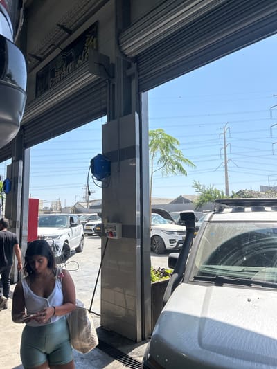 A woman in a white tank top and green shorts stands next to a car wash station, holding a phone. Several vehicles are parked in the background, including a white Land Rover. Overhead, utility poles stretch into the distance against a clear sky. Part of a building's awning and support structure frames the scene. A blue hose reel is mounted on the tiled wall of the car wash, near a power outlet.