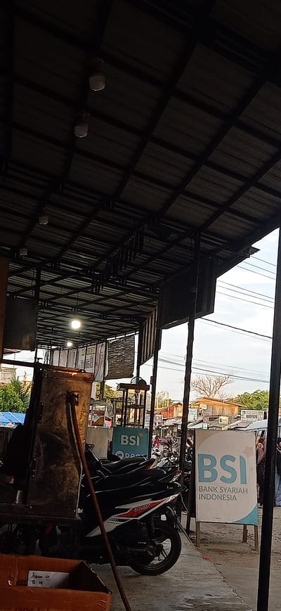 The image depicts an outdoor scene, likely a market or a street vendor area, under a covered structure with a corrugated metal roof supported by metal beams. The time of day appears to be late afternoon or early evening, with the sky visible through gaps in the roof and overhead wires suggesting an urban or semi-urban environment.
The primary subjects include several parked motorcycles, a cart with a large, dark container, and a few individuals visible in the background, suggesting ongoing activity. The setting is a street lined with buildings and shops, with more vehicles and people moving about in the distance, hinting at a bustling local scene.
Notable details include several light fixtures attached to the ceiling of the covered area. There are signs visible in the image. One prominent sign is for "BSI BANK SYARIAH INDONESIA," with the BSI logo in light blue. Another smaller sign shows "BSI" as well. There is also a sign with text that is partially visible and difficult to read clearly, but it appears to be in Indonesian. A brown cardboard box is in the foreground with some handwritten text that is also unclear. The overall mood is one of everyday life and commerce. The weather appears to be clear, with the sky a pale blue. The location is identified as Gampongteungoh, Indonesia.