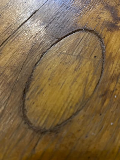 This media file shows a close-up, overhead view of a damaged wooden surface, likely a table or floor. The wood has a warm, natural tone with visible grain patterns and some darker staining or aging. An oval-shaped gouge or depression is prominently featured in the center of the frame, its edges appearing rough and worn. The surrounding wood also shows some fine scratches and imperfections. There are a few small dark specks scattered across the surface, which could be dirt or natural imperfections in the wood. The lighting appears to be artificial, casting a soft glow on the textured surface. No people, text, or specific environmental cues are visible to indicate time of day, weather, or a precise location beyond it being an interior wooden surface. The context provided, "Krasnadvorski Sel'ski Sovet, Belarus," suggests a rural setting within Belarus.
