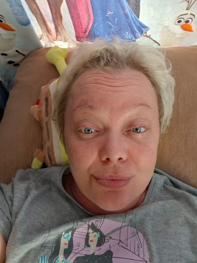 A close-up, overhead shot shows a person with fair skin and short, light blonde hair. The person is lying down, looking directly at the camera. Their eyes are open and appear to be light blue. They have visible wrinkles on their forehead and around their eyes. The person is wearing a grey t-shirt with a graphic print of two cartoon-like women. A stuffed toy, possibly a Spongebob Squarepants character, is visible next to their head. In the background, a blanket or sheet with animated characters, including what appears to be Olaf from Frozen, is visible. The setting appears to be indoors, possibly a bed or sofa. No specific location in Novoilinsky, Russia can be determined from the image.