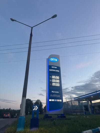 This image depicts a view of a gas station in the city of Vereya, Russia, during dusk. A prominent blue and white gas price display tower is visible on the right, showing prices for different fuel types, with "ОПТИ" and "ГАЗПРОМ НЕФТИ" branding. Below the fuel prices, "AdBlue" is advertised. To the left of the price display, a tall concrete utility pole with a street lamp stands, casting a shadow on the surrounding grass. A dark-colored car is seen driving away on a road in the background on the left. The sky is a soft blue with scattered clouds, and a crescent moon is visible. Further to the right, a portion of the gas station's canopy is visible, illuminated by blue lights. The overall environment appears to be a semi-urban or rural roadside setting.