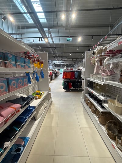 The image depicts the interior of a retail store, likely a department store or a clothing and home goods store. The scene is viewed from a low angle, looking down a wide aisle lined with shelves on either side, filled with merchandise.
On the left, shelves display items such as packaged inflatable rings, towels in various colors (pink, blue, and patterned), and what appear to be beach bags. Some of these items are also seen in wire baskets.
On the right, the shelves are stocked with home decor items. There are decorative vases, candle holders, and numerous woven baskets of different sizes. Further down the aisle, racks of clothing are visible, with a variety of garments in different colors.
The store has a modern industrial aesthetic, with a high ceiling featuring exposed beams, ductwork, and lighting fixtures. Skylights are visible in the ceiling, letting in natural light. An illuminated green exit sign with an arrow is affixed to a beam overhead.
The flooring is tiled in a light, neutral color. The overall impression is one of an organized and well-stocked retail environment. There are no individuals clearly visible in the image, but the setup implies customer traffic.
Regarding the location in the city of Skutare, Bulgaria, the provided image does not contain any explicit visual cues or contextual information to confirm this specific location.