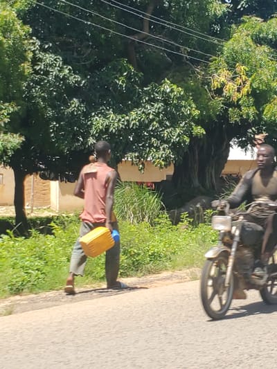 In Panyam, Nigeria, a person walks along a paved road carrying a yellow jerrycan with a blue lid. The person is wearing a reddish-orange sleeveless shirt and grey pants. To the left of the road is lush green vegetation and trees, with some power lines visible. In the background, a motorcycle is parked on the side of the road, with another person seated on it. The scene appears to be set during the daytime, with bright sunlight casting shadows. The overall atmosphere suggests a common daily activity of transporting water or other goods in a rural Nigerian setting.